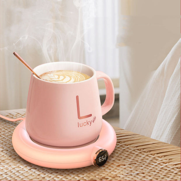 Pink mug on a cup warmer pad displaying 65°C, with steam rising and a cozy background, ideal for warm beverages.